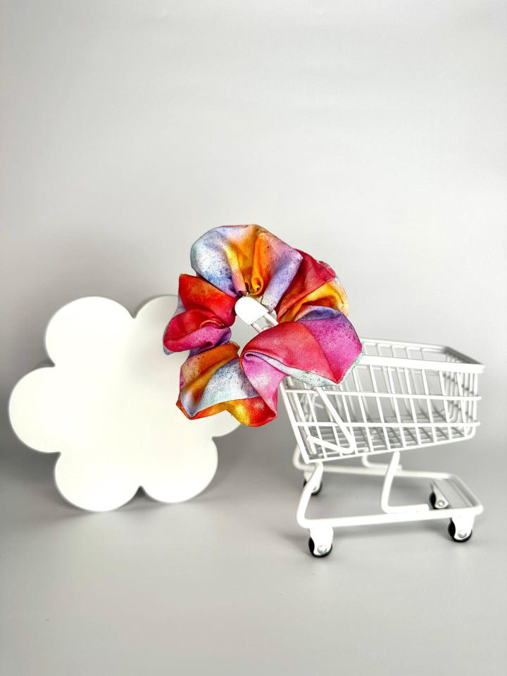 A tie dye scrunchie with a top angle view, showing the splashes of pinks, oranges, purples, blues, greens and reds with some splat-like sots on the fabric.